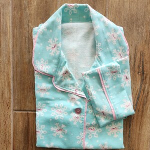 May include: A folded pajama set featuring a light blue fabric with a snowflake pattern. The snowflakes have smiling faces and pink accents. The pajamas have pink trim and a single button closure.