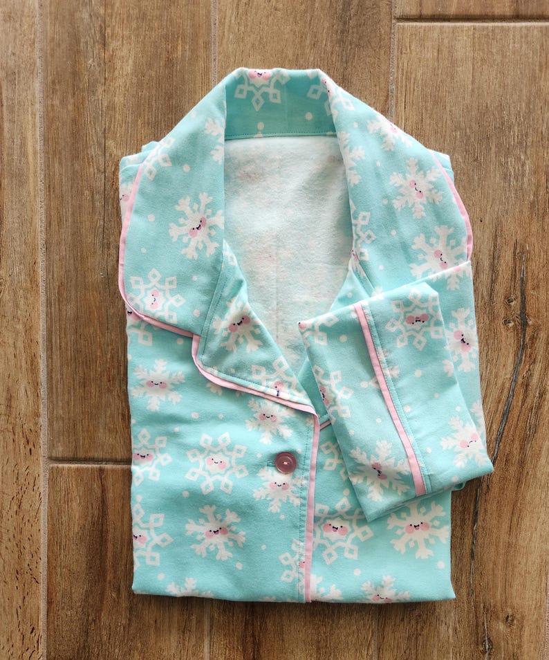 May include: A folded pajama set featuring a light blue fabric with a snowflake pattern. The snowflakes have smiling faces and pink accents. The pajamas have pink trim and a single button closure.