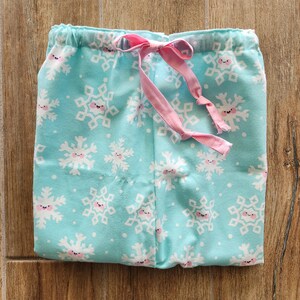 May include: Light blue pajama pants with a repeating pattern of smiling snowflakes in white and pink. The pants have a pink drawstring and an elastic waistband. The fabric is soft and appears to be cotton.