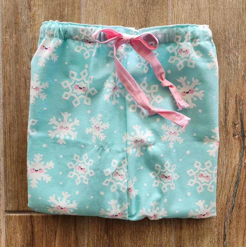 May include: Light blue pajama pants with a repeating pattern of smiling snowflakes in white and pink. The pants have a pink drawstring and an elastic waistband. The fabric is soft and appears to be cotton.
