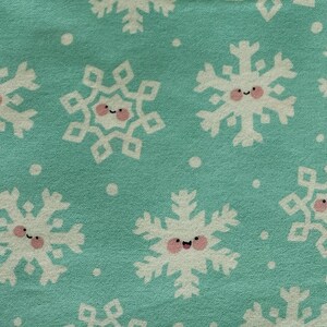 May include: A close-up of a teal fabric with a repeating pattern of white snowflakes. Each snowflake has a smiling face with pink cheeks and a small black curved mouth. Small white dots are scattered between the snowflakes.