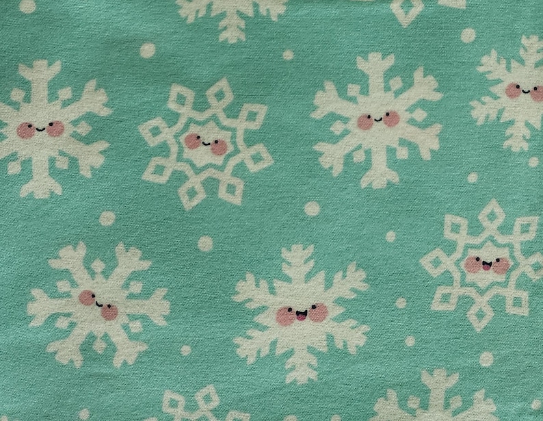 May include: A close-up of a teal fabric with a repeating pattern of white snowflakes. Each snowflake has a smiling face with pink cheeks and a small black curved mouth. Small white dots are scattered between the snowflakes.