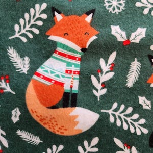 May include: A festive green fabric featuring a cartoon fox wearing a sweater with red, white, and green patterns. The design includes white snowflakes, holly, and sprigs, creating a holiday theme.