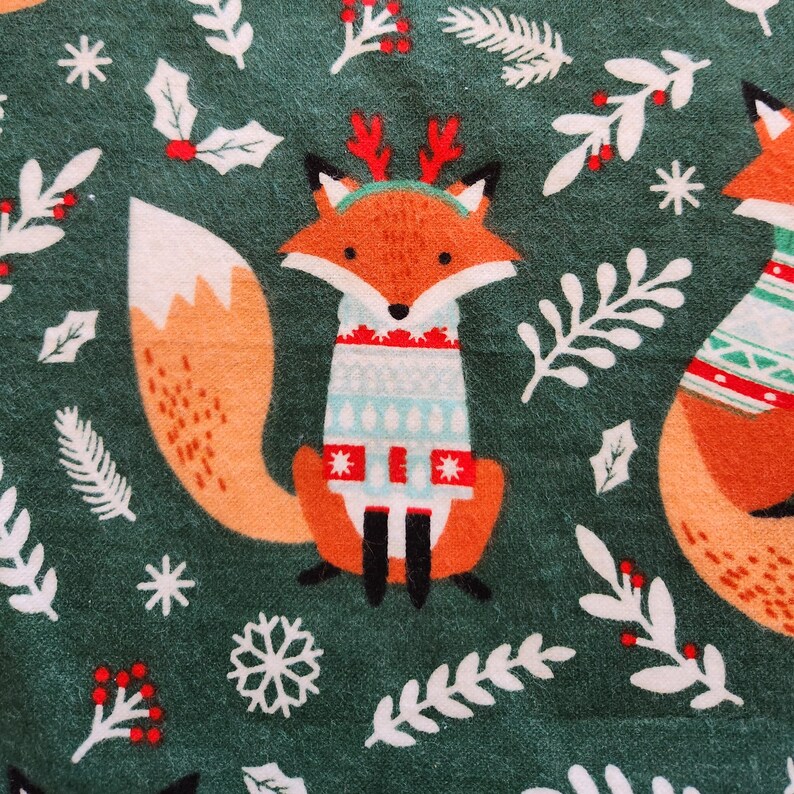 May include: A repeating pattern on a green background features cartoon foxes wearing sweaters and reindeer antlers. The foxes have orange fur, white and black accents, and are surrounded by white floral and snowflake designs. The sweaters have red, white, and blue patterns.