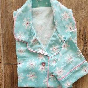 May include: A light blue pajama set with a snowflake pattern. The pajamas have a button-down top with a collar and long sleeves. The edges are trimmed with pink piping. The snowflakes have smiling faces.