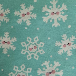 May include: Close-up of a fabric with a repeating pattern of white snowflakes with smiling faces and pink cheeks on a light blue background. Small white dots are scattered throughout the design.