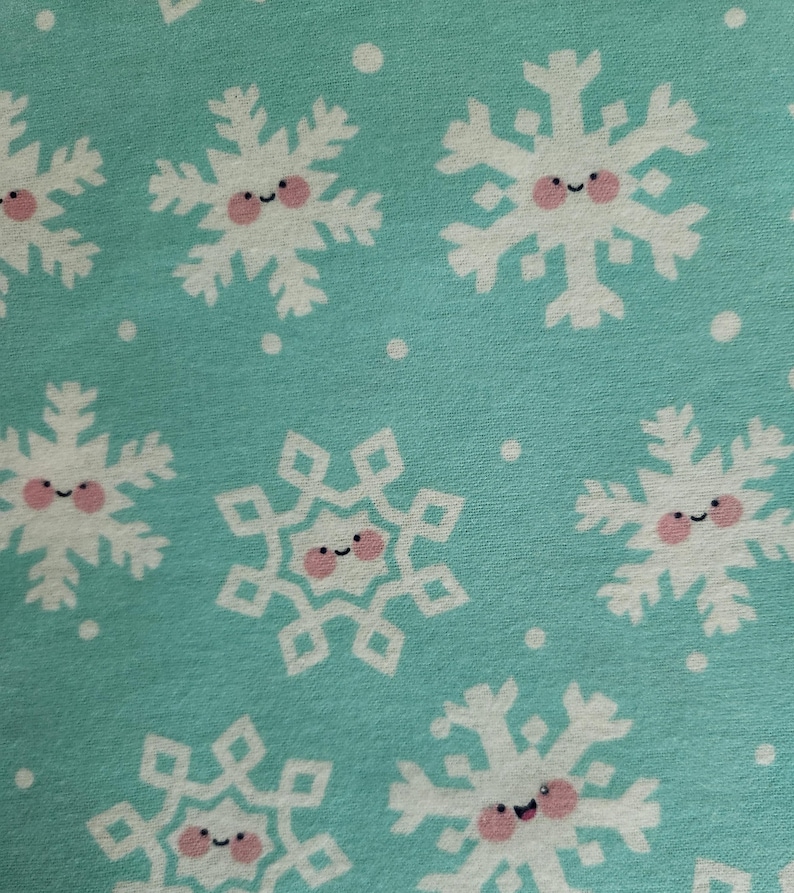 May include: Close-up of a fabric with a repeating pattern of white snowflakes with smiling faces and pink cheeks on a light blue background. Small white dots are scattered throughout the design.