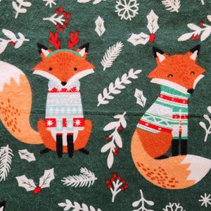 May include: A green fabric featuring two orange foxes wearing sweaters with festive patterns. The foxes are surrounded by white snowflakes, holly, and other winter foliage. The foxes have black ears and noses.