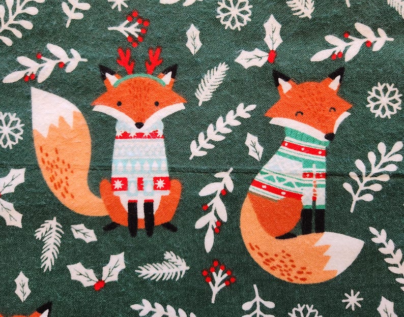 May include: A green fabric featuring two orange foxes wearing sweaters with festive patterns. The foxes are surrounded by white snowflakes, holly, and other winter foliage. The foxes have black ears and noses.