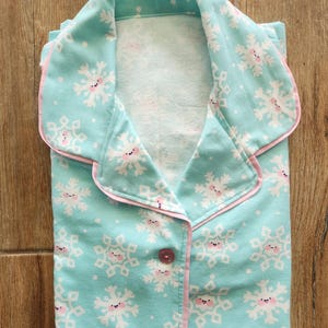 May include: Folded pajama set in a light blue color, featuring a repeating pattern of smiling snowflakes. The pajamas have pink trim around the collar, cuffs, and button placket. A single pink button is visible.