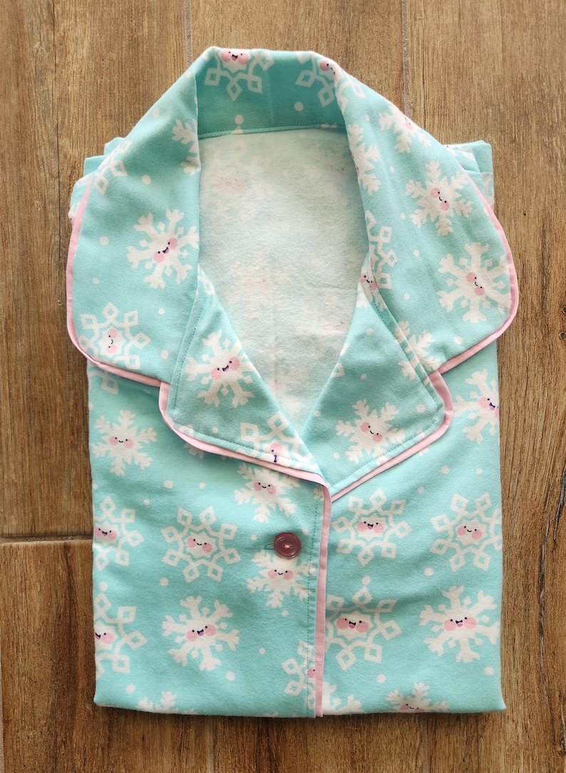 May include: Folded pajama set in a light blue color, featuring a repeating pattern of smiling snowflakes. The pajamas have pink trim around the collar, cuffs, and button placket. A single pink button is visible.
