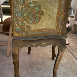 May include: A side table with curved legs featuring a gold and teal finish. The side panel displays an ornate, raised scrollwork pattern in gold against a teal background, framed by a border of small teal dots on a gold surface with a distressed texture.