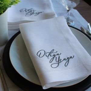May include: A crisp white cloth napkin folded neatly on a plate, featuring the embroidered dark gray cursive text "Ortega Ycaza" in the corner. The napkin has a stitched hem border and is displayed alongside a fork and a glass.
