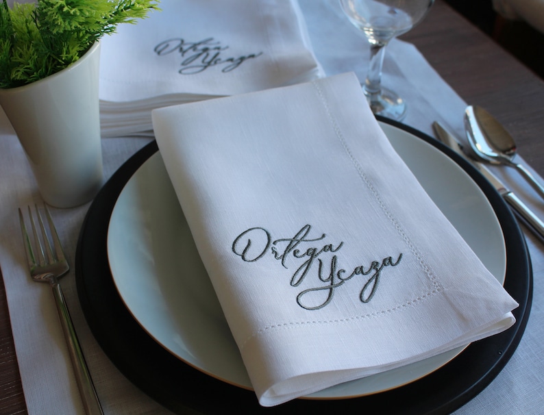 May include: A crisp white cloth napkin folded neatly on a plate, featuring the embroidered dark gray cursive text "Ortega Ycaza" in the corner. The napkin has a stitched hem border and is displayed alongside a fork and a glass.