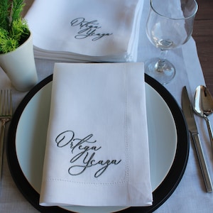 May include: A white fabric napkin featuring dark grey embroidered cursive text that reads Ortega Ycaza. The napkin is folded neatly on a white plate with a black rim, accompanied by a silver fork, a silver knife, and a silver spoon.