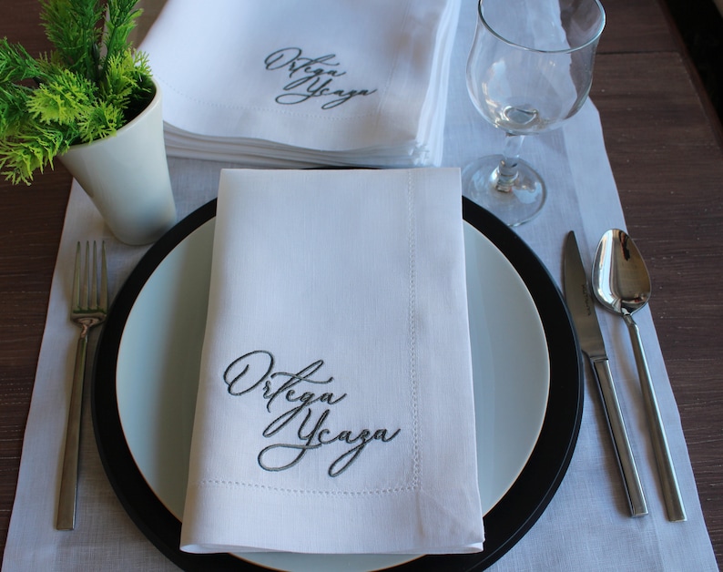 May include: A white fabric napkin featuring dark grey embroidered cursive text that reads Ortega Ycaza. The napkin is folded neatly on a white plate with a black rim, accompanied by a silver fork, a silver knife, and a silver spoon.