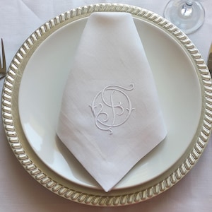 May include: A white linen napkin with a monogrammed initial "B" embroidered in the center. The napkin is folded and placed on a silver charger plate.