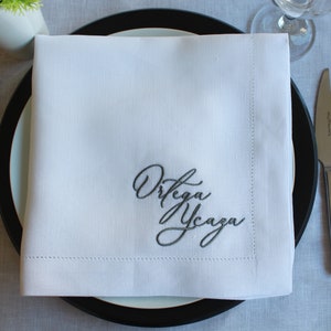 May include: A square white fabric napkin featuring a decorative hemstitch border and dark gray embroidered cursive text that reads Ortega Ycaza, centered on a black and white dinner plate with a fork, knife, and spoon placed to the sides.
