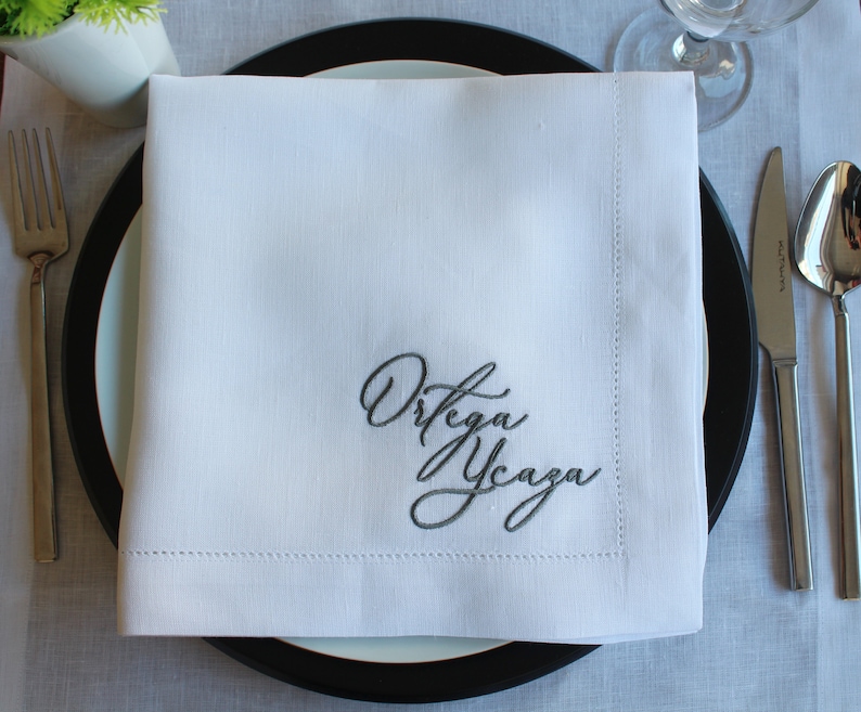 May include: A square white fabric napkin featuring a decorative hemstitch border and dark gray embroidered cursive text that reads Ortega Ycaza, centered on a black and white dinner plate with a fork, knife, and spoon placed to the sides.