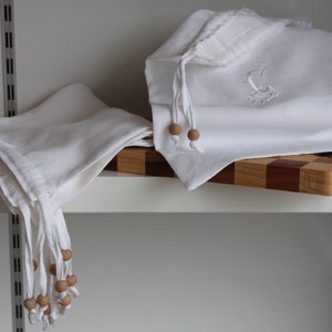 May include: Two white linen bags with wooden bead ties. One bag has an embroidered letter C. The bags are on a wooden cutting board with a brown leather cord and colorful beads.