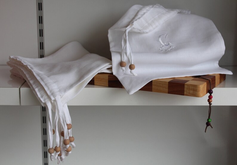 May include: Two white linen bags with wooden bead ties. One bag has an embroidered letter C. The bags are on a wooden cutting board with a brown leather cord and colorful beads.