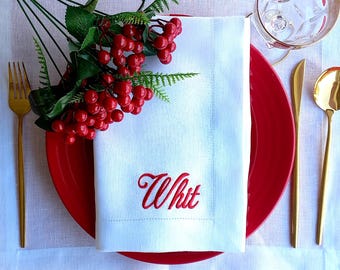 May include: A white linen napkin featuring the embroidered red script name Whit, placed on a solid red plate. The setting includes gold-toned cutlery, a glass, and a decorative sprig of red berries with green leaves resting on the folded napkin.