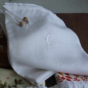 May include: White linen drawstring bag with wooden beads and an embroidered letter "C" with a crown above it.