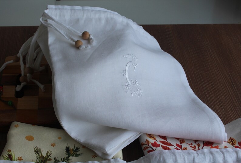 May include: White linen drawstring bag with wooden beads and an embroidered letter "C" with a crown above it.