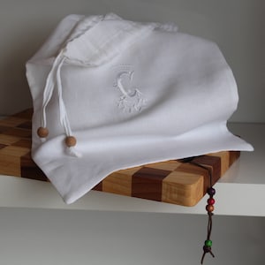 May include: White linen bread bag with a wooden bead drawstring and an embroidered letter C. The bag is sitting on a wooden cutting board with a brown leather cord with colorful beads hanging off the edge.