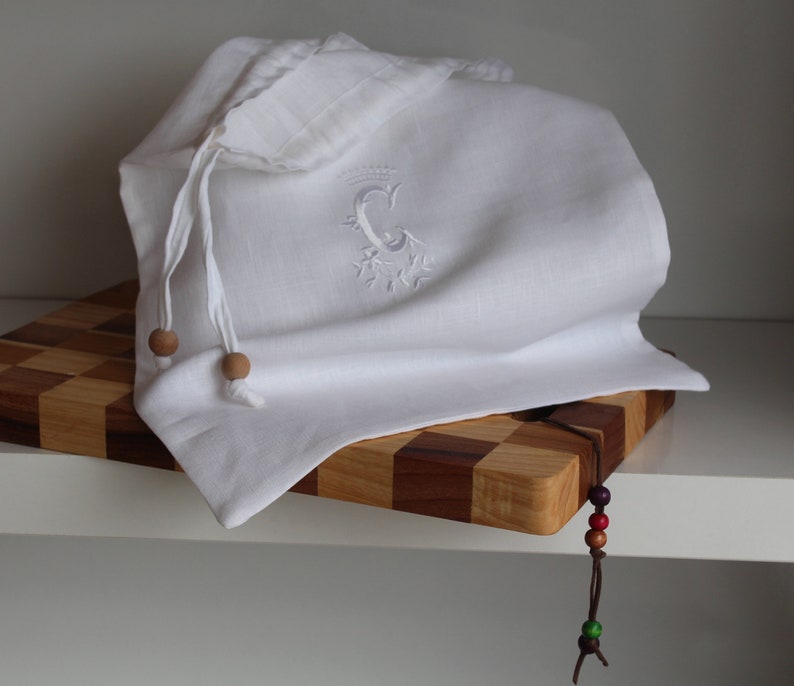 May include: White linen bread bag with a wooden bead drawstring and an embroidered letter C. The bag is sitting on a wooden cutting board with a brown leather cord with colorful beads hanging off the edge.