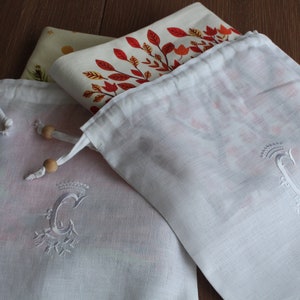 May include: Two white cotton drawstring bags with wooden toggles. Both bags have a white embroidered letter "C" with a crown above the letter. The bags are folded and stacked on top of a patterned fabric.
