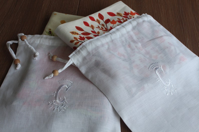 May include: Two white cotton drawstring bags with wooden toggles. Both bags have a white embroidered letter "C" with a crown above the letter. The bags are folded and stacked on top of a patterned fabric.