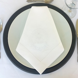 Peut inclure: Serviette de table en lin blanc avec une bordure noire, pliée en forme de triangle. La serviette est ornée d'un motif brodé représentant un cercle avec une ancre stylisée à l'intérieur.