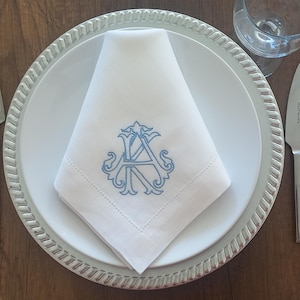 Könnte beinhalten: Eine weiße Leinen-Serviette mit einem blauen gestickten Monogramm der Buchstaben "AK" auf einem weißen Teller mit silbernem Rand.