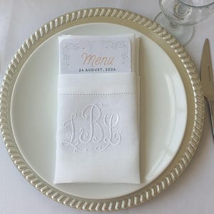 Könnte beinhalten: Eine weiße Leinen-Serviette mit einem Monogramm "J.B.L." in weißer Stickerei. Die Serviette ist gefaltet und auf einem silbernen Untersetzer mit einem weißen Teller platziert. Eine kleine Karte mit dem Text "Menu 24 August, 2024" ist in die Serviette gesteckt.