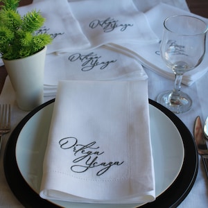 May include: A set of white cloth napkins featuring the embroidered black text Ortega Ycaza in a cursive script. The napkins are folded and arranged on a table setting with a white plate, a black charger, a fork, a spoon, a knife, and a glass.