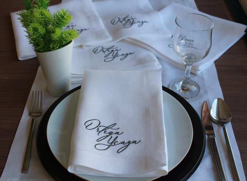 May include: A set of white cloth napkins featuring the embroidered black text Ortega Ycaza in a cursive script. The napkins are folded and arranged on a table setting with a white plate, a black charger, a fork, a spoon, a knife, and a glass.