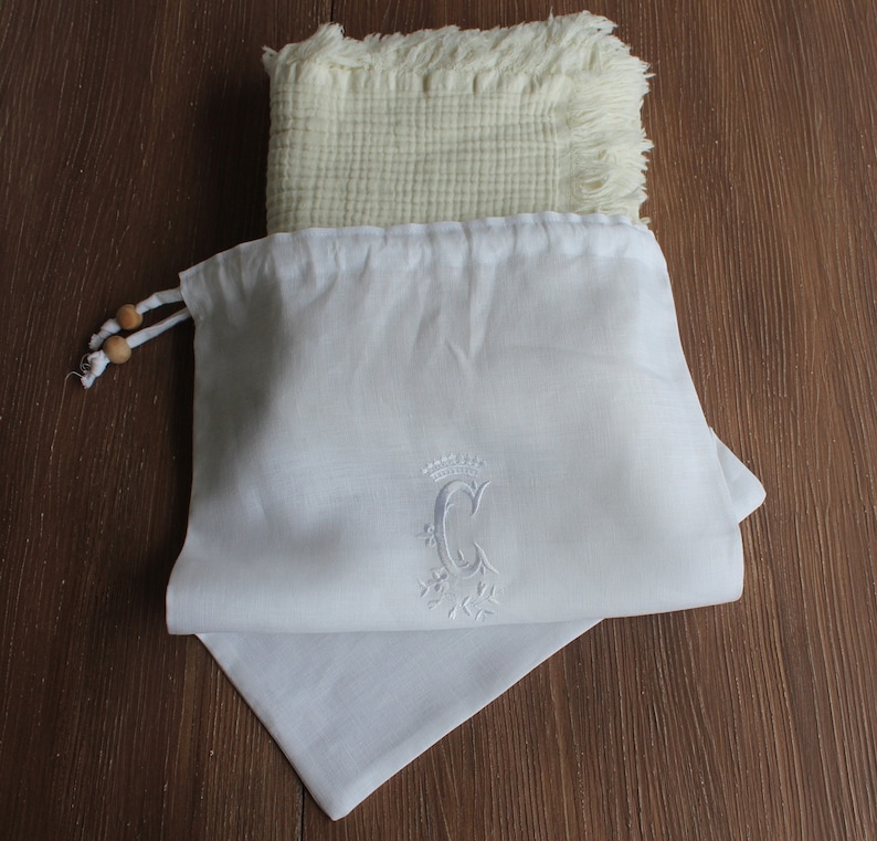 May include: White linen drawstring bag with a white embroidered letter "C" with a crown and floral design.