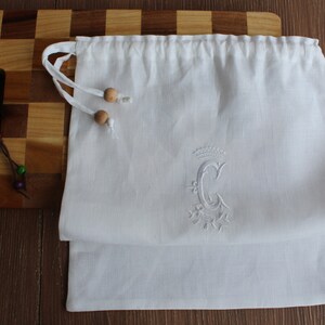 May include: White linen drawstring bag with a white embroidered letter "C" with a crown above it and a floral design below it. The bag has wooden toggles.