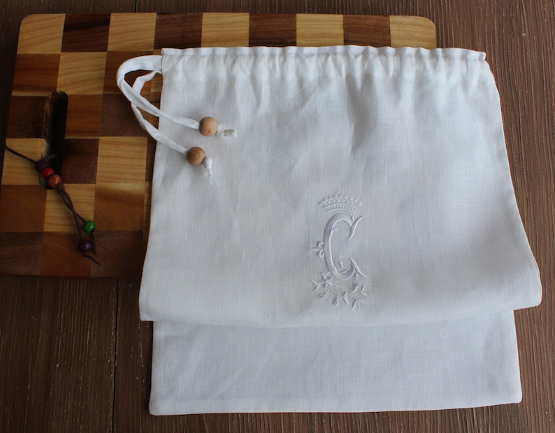May include: White linen drawstring bag with a white embroidered letter "C" with a crown above it and a floral design below it. The bag has wooden toggles.