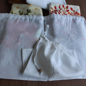 May include: Three white cotton drawstring bags with a monogrammed letter C embroidered on each. The bags are folded and arranged on a wooden surface.