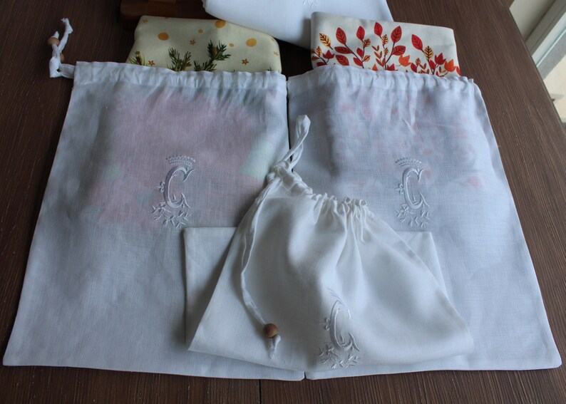 May include: Three white cotton drawstring bags with a monogrammed letter C embroidered on each. The bags are folded and arranged on a wooden surface.