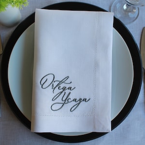 May include: A crisp white rectangular cloth napkin featuring dark grey embroidered cursive text that reads Ortega Ycaza. The napkin is folded neatly and placed centrally on a round plate with a wide black rim and a light blue center.