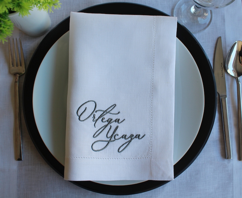 May include: A crisp white rectangular cloth napkin featuring dark grey embroidered cursive text that reads Ortega Ycaza. The napkin is folded neatly and placed centrally on a round plate with a wide black rim and a light blue center.