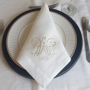 May include: A white linen napkin with a monogram of the letters "A" and "A" and the year "1989" embroidered in white thread. The napkin is folded and placed on a black and white striped plate.