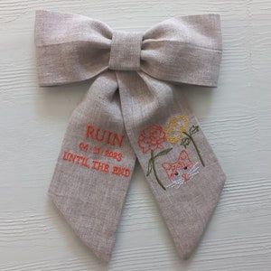 May include: A beige linen bow with embroidered text that reads "RUIN 04.11.2023 UNTIL THE END" and a small embroidered orange cat with green flowers.