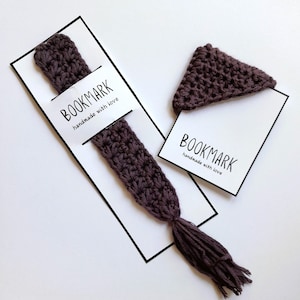 May include: Two crocheted bookmarks, one with a tassel and the other with a triangular shape. Both bookmarks are a dark purple color and have a white tag attached that says "BOOKMARK handmade with love".