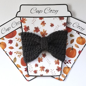 May include: A gray knitted bow tie on a white background with a pattern of orange pumpkins and fall leaves. The background is a paper cutout of a coffee cup with the text "Cup Cozy" on it.