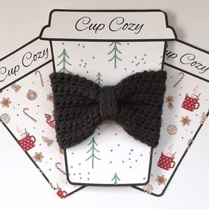 Peut inclure: Un protège-tasse en forme de nœud papillon tricoté gris foncé est posé sur une carte en forme de gobelet en papier. La carte présente un motif sur le thème de l'hiver avec des arbres, des flocons de neige et des tasses. Les mots "Cup Cozy" sont imprimés en haut.
