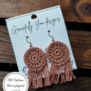 Puede incluir: Un par de pendientes de ganchillo hechos a mano en un tono cálido y terroso. Los pendientes presentan un diseño circular con un patrón calado central y largas borlas. Se muestran en una tarjeta con el texto "Gracefully You designs" y "PDF Pattern Not a physical product".
